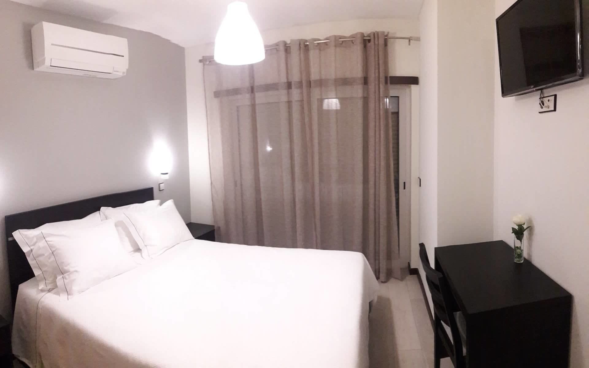 Et hotelværelse med seng, skrivebord, stol, aircondition, fjernsyn og gardin.