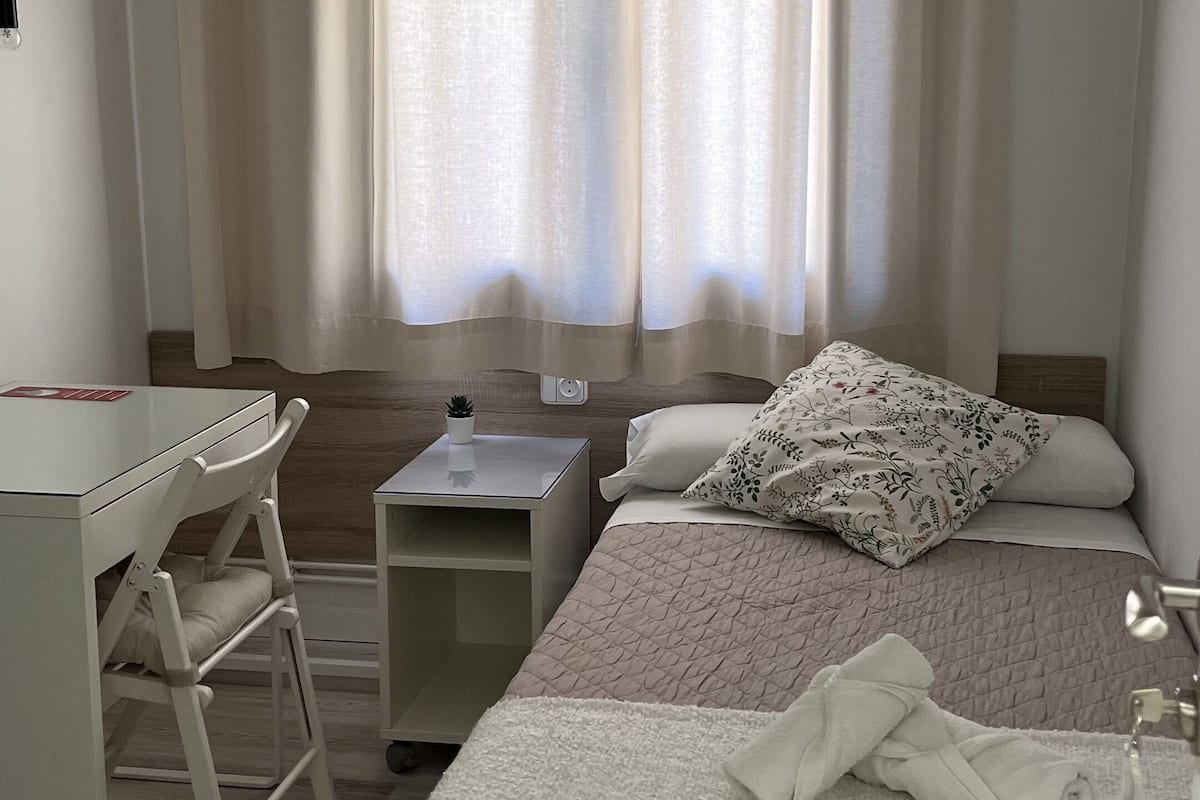 Une chambre à coucher comprenant un lit, un bureau, une chaise, une table de chevet et une fenêtre avec des rideaux.