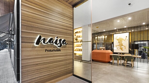 A modern hotel lobby with a wooden reception desk, a glass partition, and a seating area with chairs and a table. - Protur Naisa Palma Hotel (Palma de Mallorca)