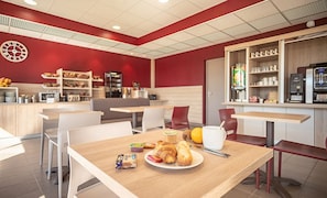 Um café com esquema de cores vermelho e branco, com mesas que exibem itens de café da manhã como croissants e suco, e uma máquina de café ao fundo.