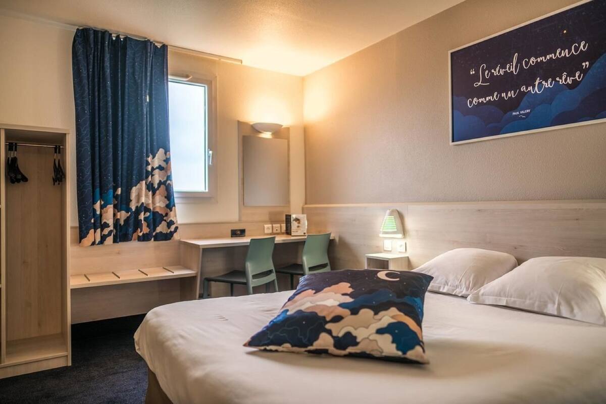 A hotel room with a bed, a desk, a chair, a wardrobe, and a wall with a quote.