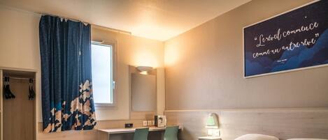 A hotel room with a bed, a desk, a chair, a wardrobe, and a wall with a quote.