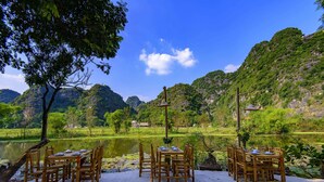 A dining table set outdoors with chairs, surrounded by a natural landscape with mountains and a body of water. - Trang An Bungalow (Tay Hoa Lu)