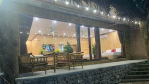 A cozy outdoor seating area with wooden furniture and a stone wall, overlooking a well-lit interior space with large windows. - Trang An Bungalow (Tay Hoa Lu)
