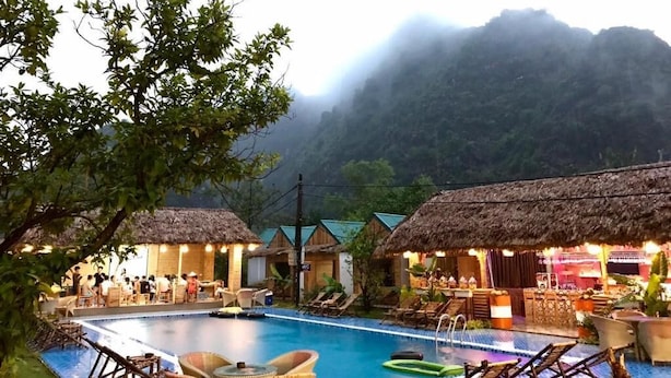 A pool area with lounge chairs and thatched-roof huts, surrounded by lush greenery and misty mountains. - Trang An Bungalow (Tay Hoa Lu)