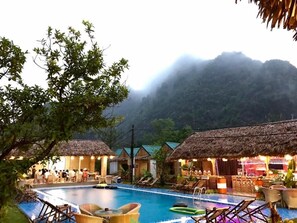 A pool area with lounge chairs and thatched-roof huts, surrounded by lush greenery and misty mountains. - Trang An Bungalow (Tay Hoa Lu)