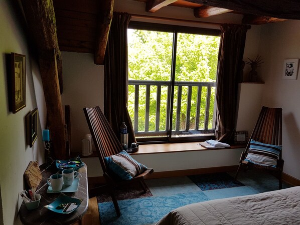 Una habitación con un ventanal grande, dos sillas, una cama, una mesita con tazas y un hervidor, y una alfombra estampada.