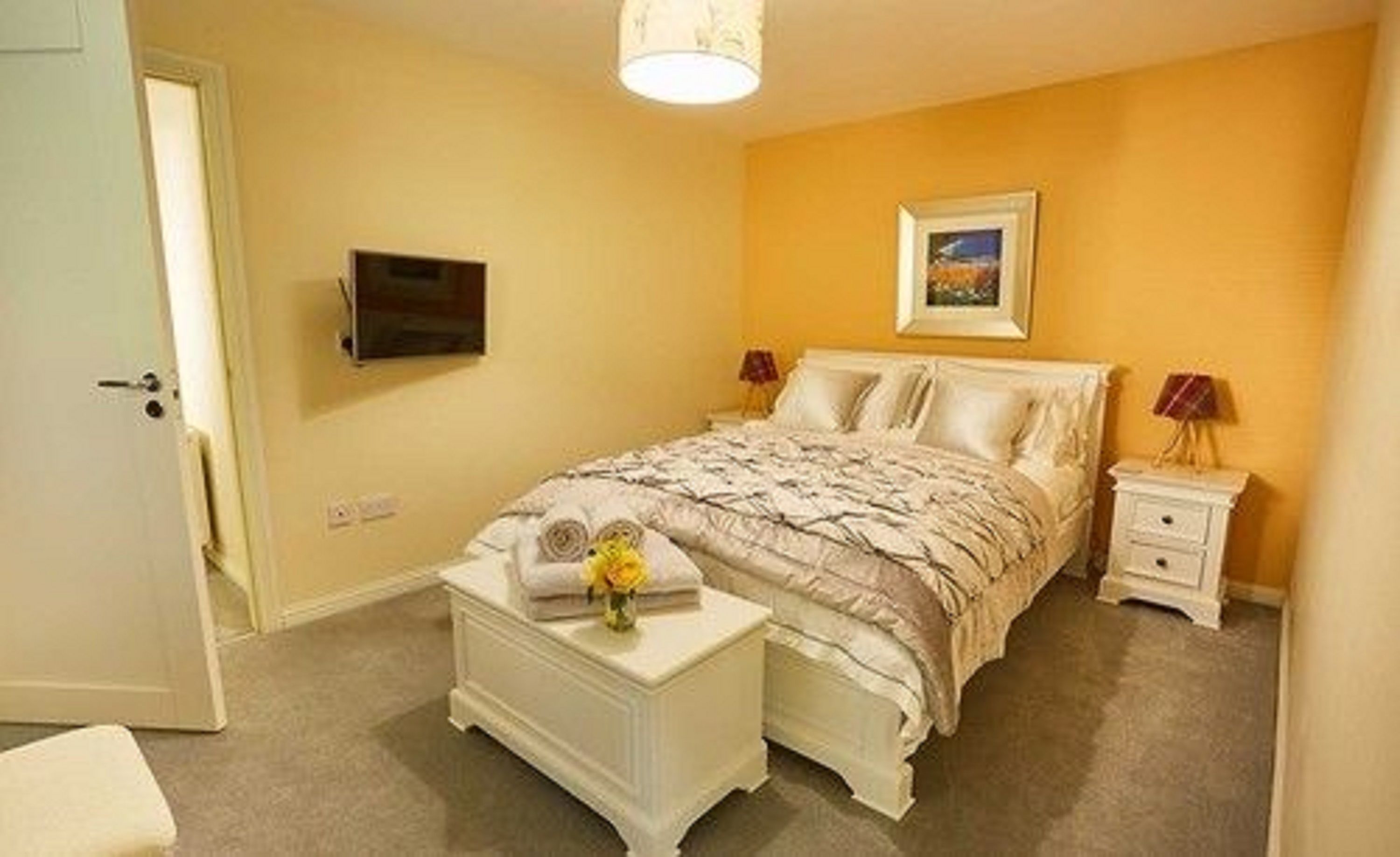 A bedroom with a bed, a TV mounted on the wall, a bedside table with a lamp, and a small table with a vase of flowers.