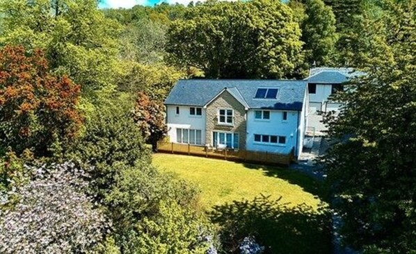 Une maison à deux étages avec une grande pelouse à l’avant, entourée d’arbres denses.