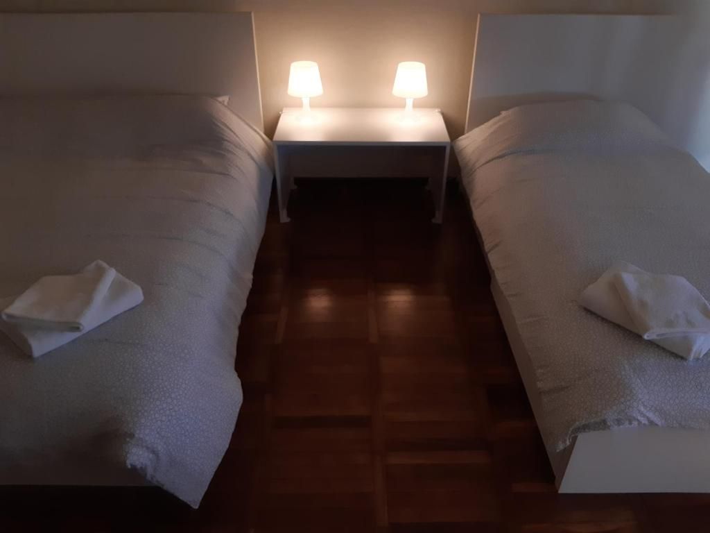 Habitación de hotel con dos camas, cada una con una almohada y una toalla doblada. Hay una mesita de noche con dos lámparas entre las camas.