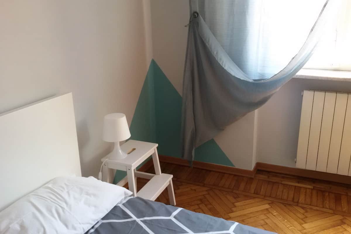 A bedroom with a bed, a nightstand with a lamp, a window with curtains, and a radiator.