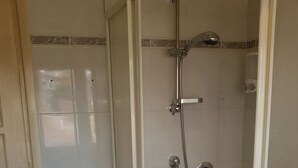 Une douche équipée d’un pommeau de douche à main et d’un robinet mural, d’une petite poubelle et d’une serviette blanche accrochée au mur.