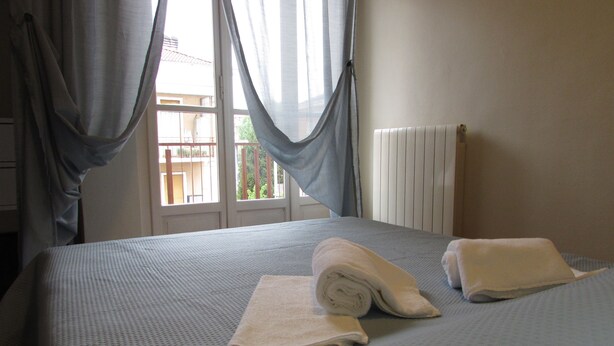 Un lit bien fait, avec une serviette pliée et une autre enroulée, une fenêtre avec des rideaux et un radiateur.