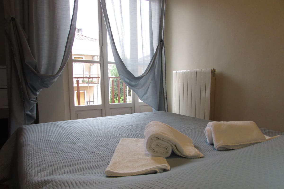 A neatly made bed with a folded towel and a rolled towel, a window with curtains, and a radiator.