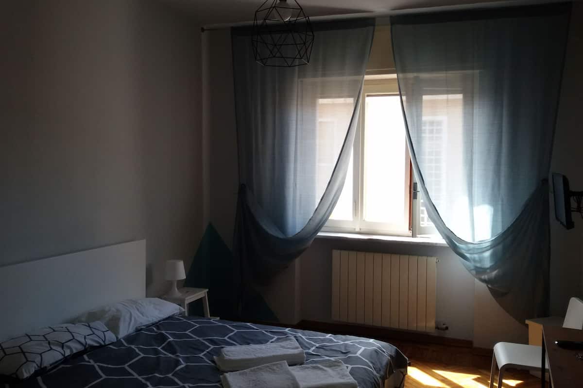 A bedroom with a bed, a bedside table with a lamp, a chair, and a window with sheer curtains.