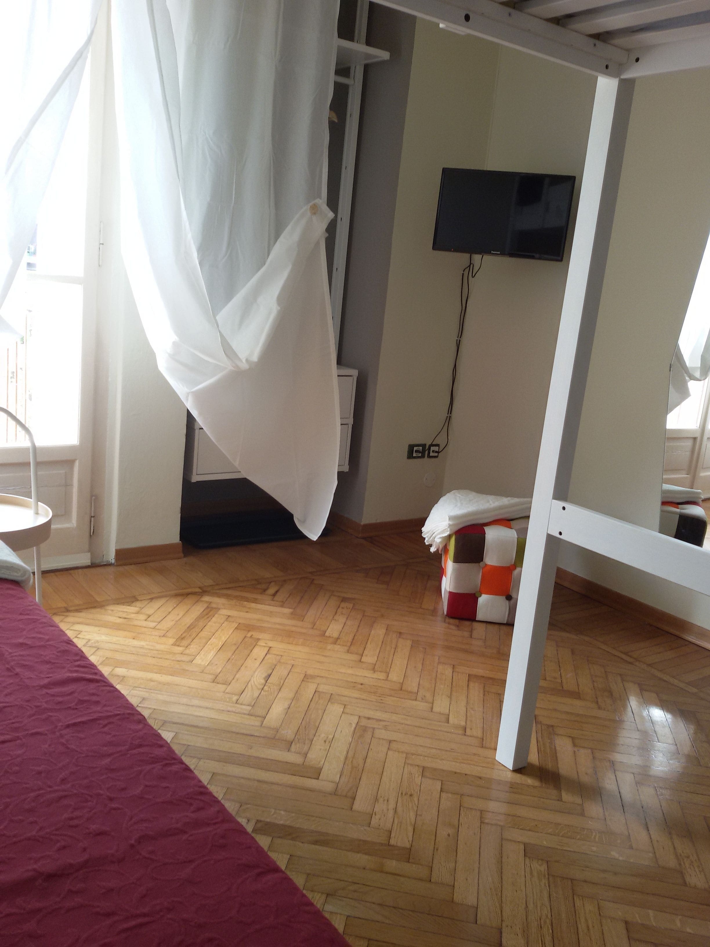 Una habitación con piso de madera, una cama con cubrecama rojo, una cortina blanca, un televisor montado en la pared y una mesita.