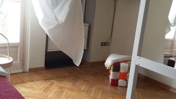 Una habitación con piso de madera, una cama con cubrecama rojo, una cortina blanca, un televisor montado en la pared y una mesita.