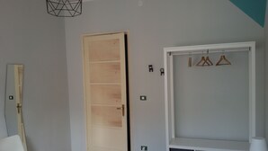 Une pièce avec une porte en bois, une chaise blanche, une armoire blanche avec des cintres et une petite table avec une lampe.