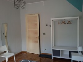 Una habitación con una puerta de madera, una silla blanca, un armario blanco con perchas y una mesita con una lámpara.