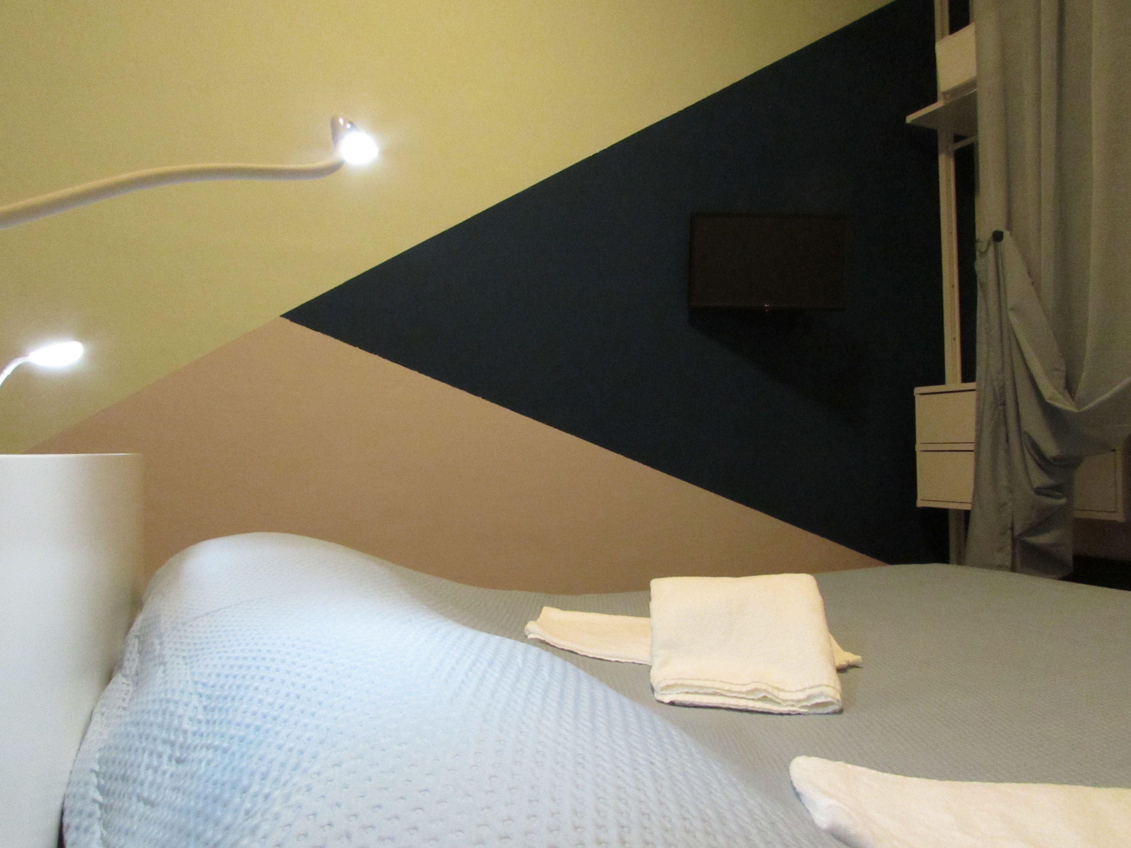 A hotel room with a bed, a bedside lamp, a flat-screen TV mounted on the wall, and a window with curtains.