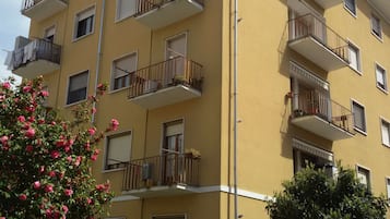 Un immeuble jaune à plusieurs étages, doté de balcons, d’une entrée fermée par un portail et d’un petit arbre aux fleurs roses.