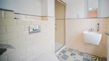 Une salle de bain avec des carreaux de céramique blancs rectangulaires, un lavabo blanc avec un miroir au-dessus, et un espace douche.