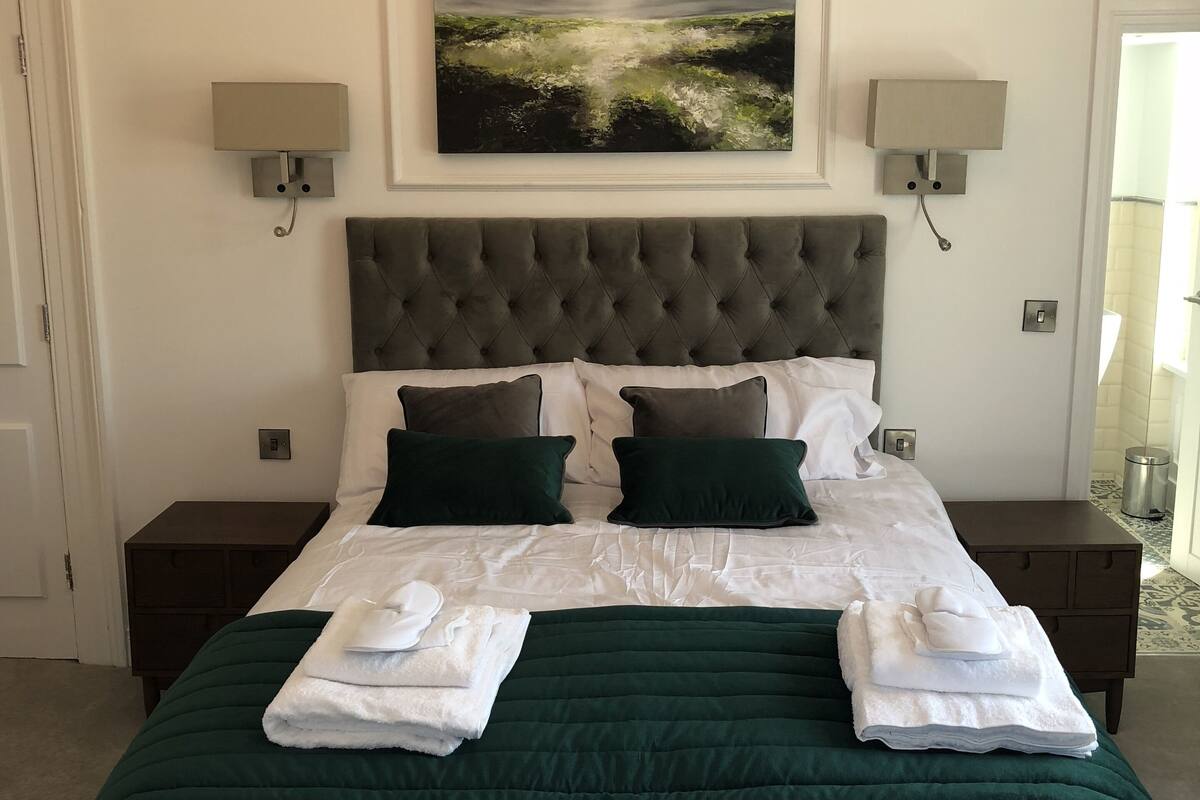 Una cama bien hecha con cubrecama verde y sábanas blancas, dos almohadas verde oscuro y dos toallas blancas dobladas sobre la cama.