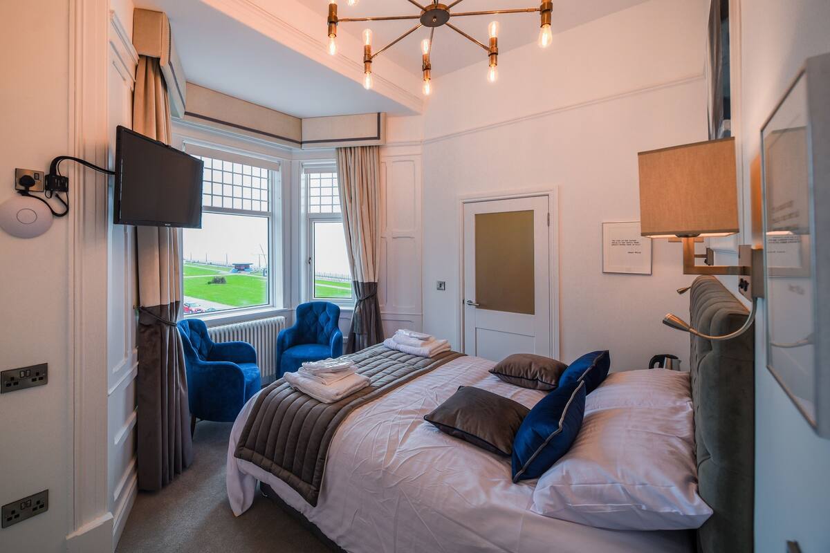 Una habitación de hotel con cama, televisión, ventana con cortinas, una silla y una lámpara.