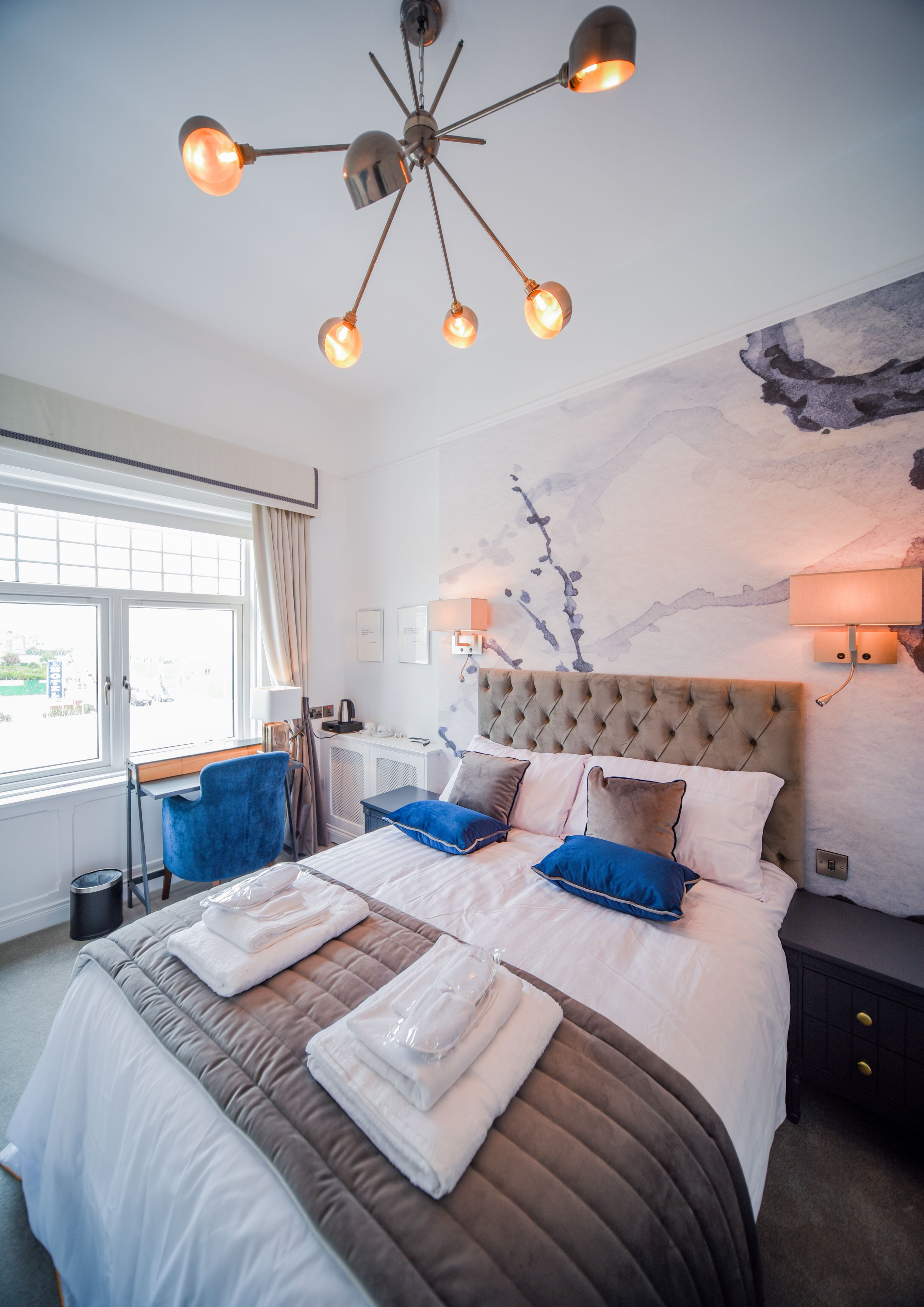 Habitación de hotel con una cama grande, un escritorio y una silla. Se ve una ventana con cortinas, una pared con un cuadro y una lámpara en el techo.