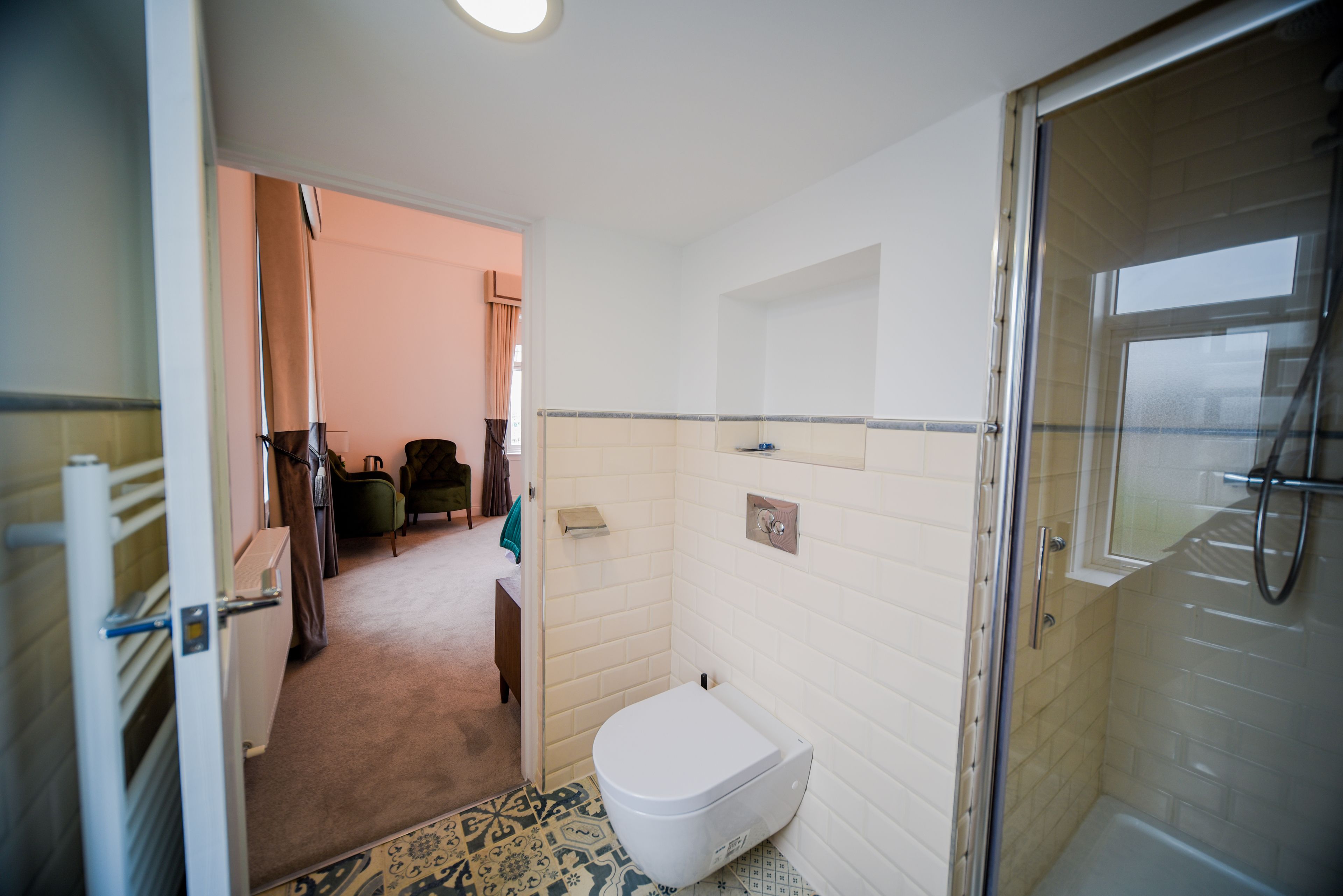Ein Badezimmer mit Toilette, Dusche und einem kleinen Waschbecken. Auf dem Boden liegt ein Teppich mit Muster und eine Tür führt in einen weiteren Raum.