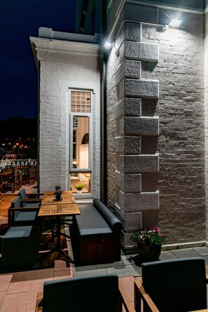 Une terrasse extérieure avec des tables et des bancs en bois, un mur de pierre et une vue sur la ville la nuit.