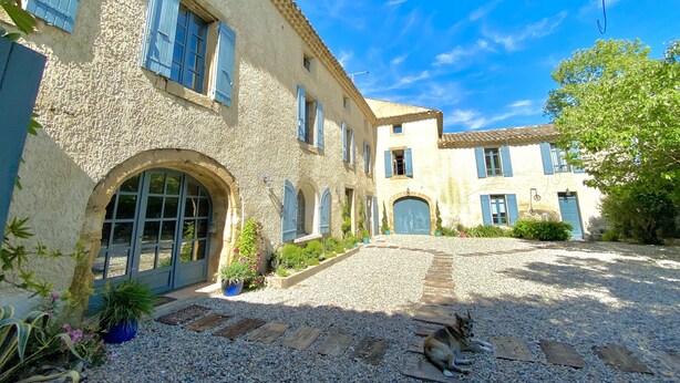 Un bâtiment rustique avec une cour en gravier, un chien allongé sur le sol et un ciel bleu dégagé.