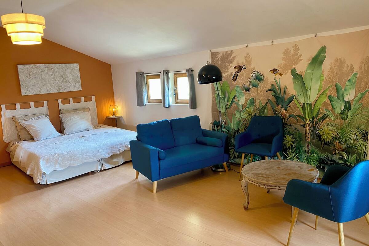 Ein Zimmer mit einem Bett, einem blauen Sofa, zwei Sesseln, einem kleinen Tisch, einer Stehlampe und einer Wand mit einem tropischen Wandbild.
