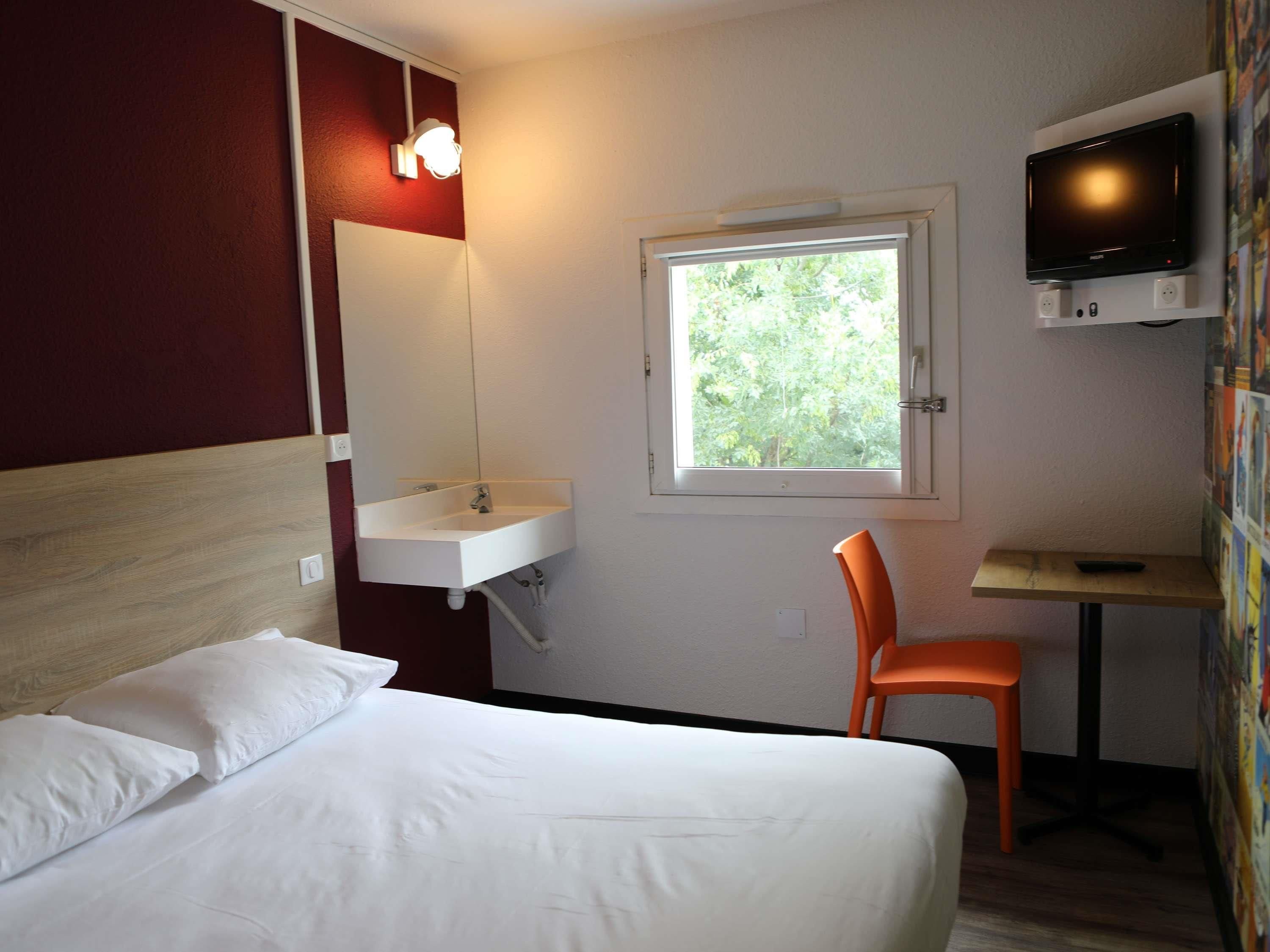 A hotel room with a bed, a white sink, a mirror, a window with a view of trees, a television, and a small table.