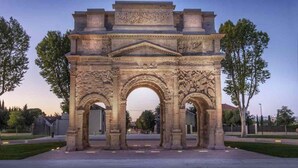 Un gran arco de piedra con intrincados grabados y un frontón central, rodeado de una zona pavimentada con árboles.