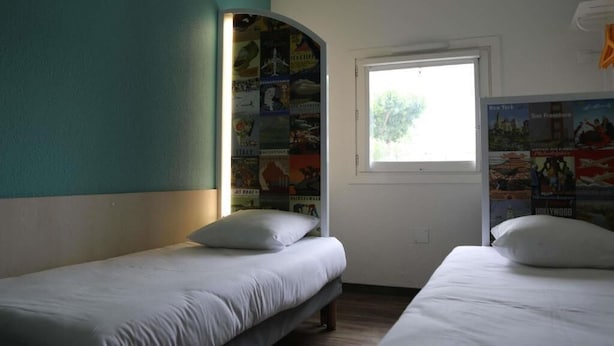 Une chambre avec deux lits simples, une fenêtre donnant sur un beau paysage et un mur décoré d’affiches de voyage.