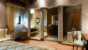 A hotel room with a bed, a sitting area with a chair and a table, a bathroom with a mirror, and a shower area. - Villa Armena (Buonconvento)