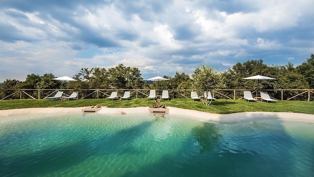 A pool area with clear water, surrounded by white lounge chairs and umbrellas, under a cloudy sky. - Villa Armena (Buonconvento)