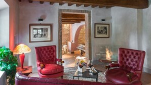 A cozy living room with red leather armchairs, a glass coffee table, and a red lamp. There are framed pictures on the walls and a stone wall in the background. - Villa Armena (Buonconvento)