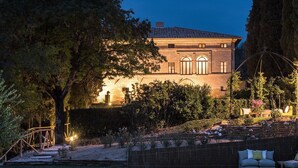 A two-story house with a brick exterior, large windows, and a well-maintained garden with various plants and flowers. - Villa Armena (Buonconvento)