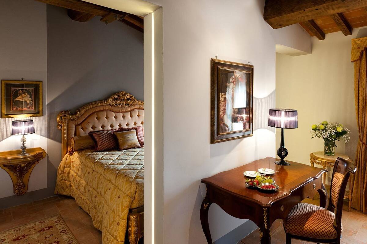 A bedroom with a large bed, a bedside table with a lamp, a framed picture on the wall, and a small table with a vase of flowers.