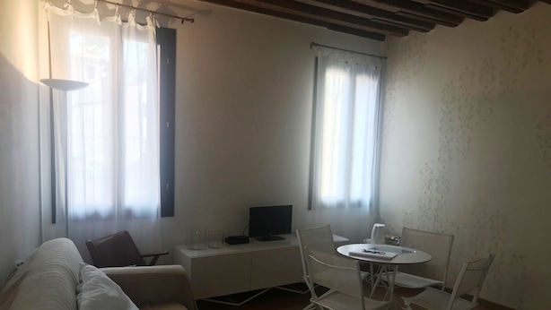 Ein Wohnzimmer mit einer Couch, einem kleinen Tisch und einem Fernseher. Zwei Fenster mit Vorhängen und eine Holzbalkendecke sind vorhanden.