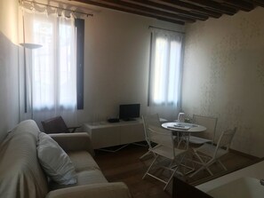 A living room with a sofa, a small table, and a TV. There are two windows with curtains, and a wooden ceiling.
