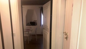 Ein enger Flur führt zu einem Essbereich mit Tisch und Stühlen, einem Küchenbereich mit Arbeitsfläche und Geräten sowie einer Tür mit Griff.