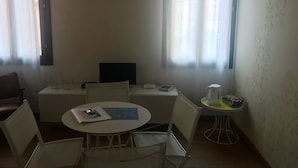 Ein kleiner, gut beleuchteter Raum mit einem runden Tisch, zwei Stühlen, einem Fernseher, einem kleinen Tisch mit einer Karaffe und Tassen sowie einer Tischkarte auf dem Tisch.
