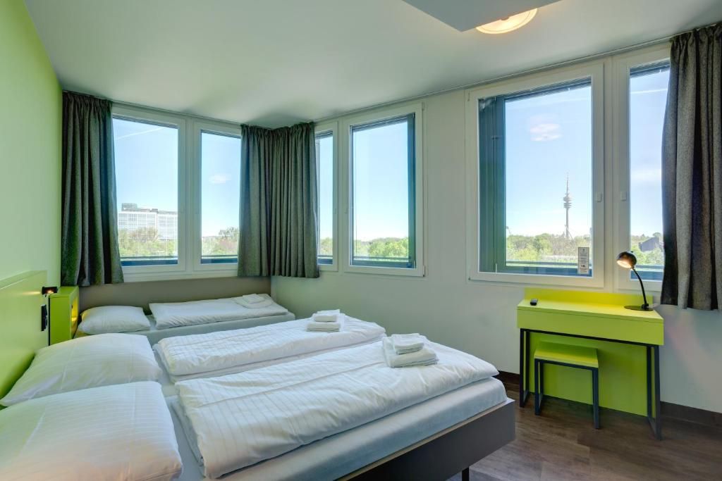 Ein Hotelzimmer mit zwei Betten, einem Schreibtisch und großen Fenstern mit Blick auf die Stadt.