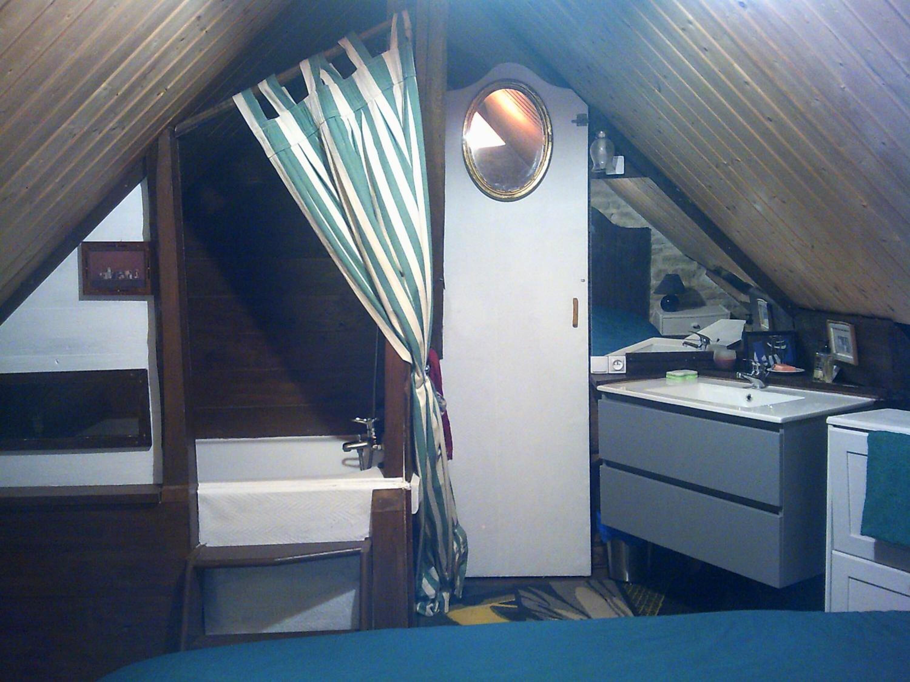 Une petite chambre mansardée étroite, avec une seule fenêtre, un miroir rond, un lavabo et un lit.