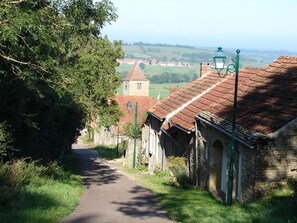 A narrow rural road flanked by stone buildings with red-tiled roofs and a church tower in the distance. - Chambres d'hôtes au verger de Marcilly (Marcilly-Ogny)