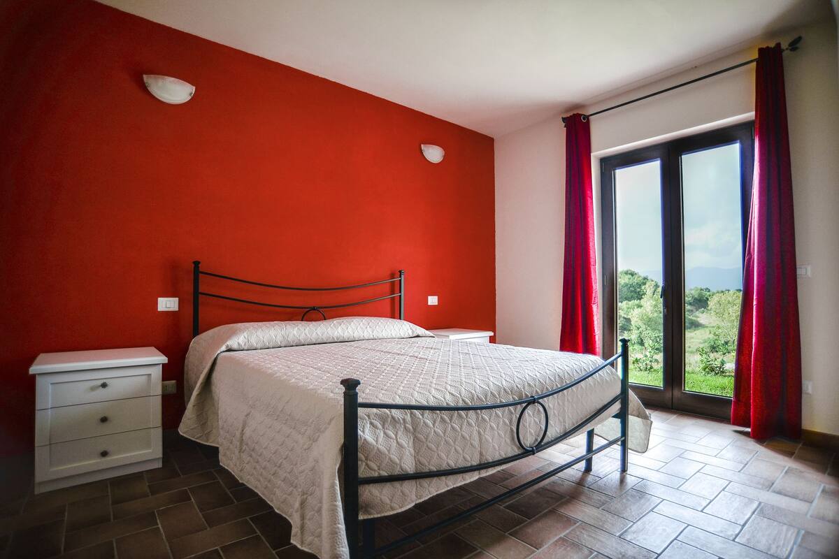 A bedroom with a bed, a nightstand, a window with curtains, and a view of greenery outside.