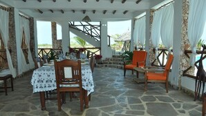 A covered outdoor dining area with stone flooring, wooden tables and chairs, and a view of the beach. - Coral Sea Villas (Malindi)